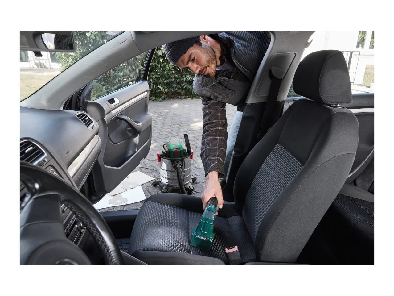 Man cleaning car interior with a wet and dry vacuum cleaner and a handheld upholstery cleaner.