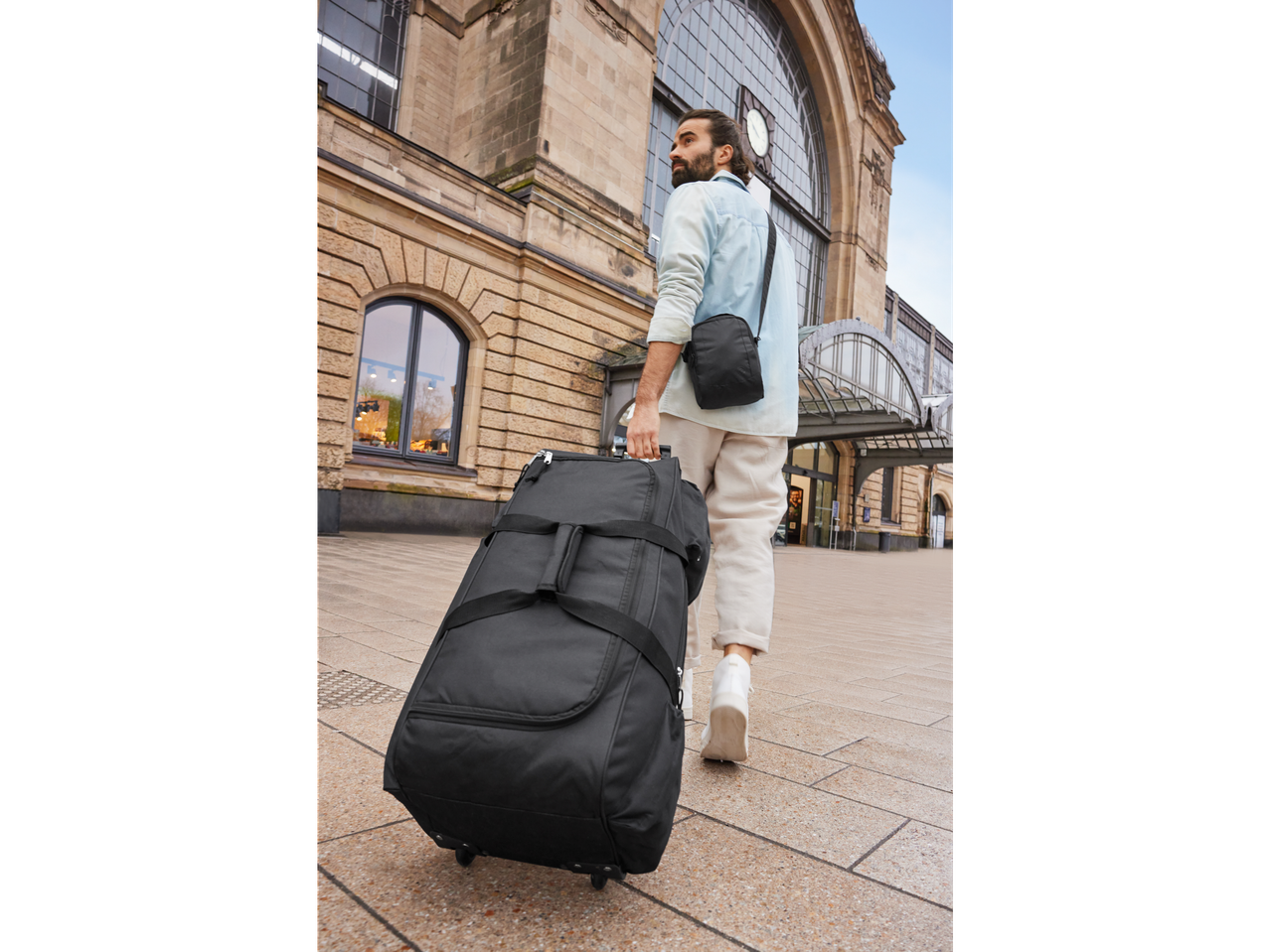 Man pulling wheeled duffel bag and shoulder bag at train station.