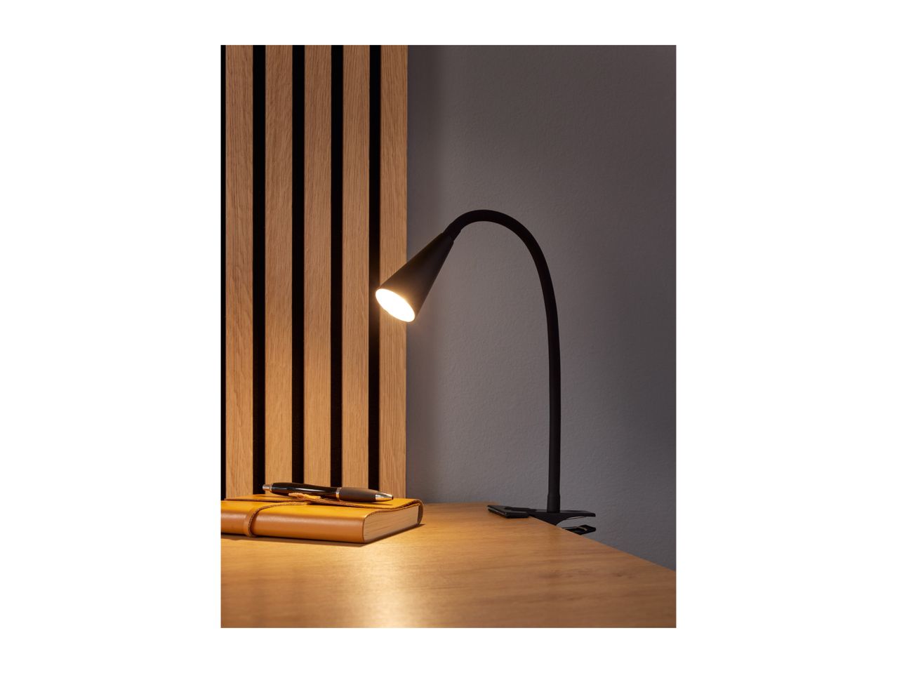 A black clamp-on desk lamp illuminates a wooden desk with a journal and pen.