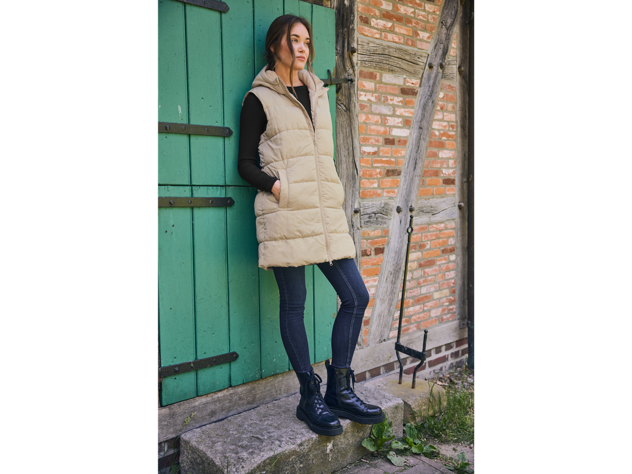 Woman in a beige puffer vest and dark skinny jeans leaning against a green door.