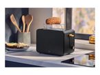 Black toaster with a bun on top, wooden utensils, and a plate of food on a kitchen counter.