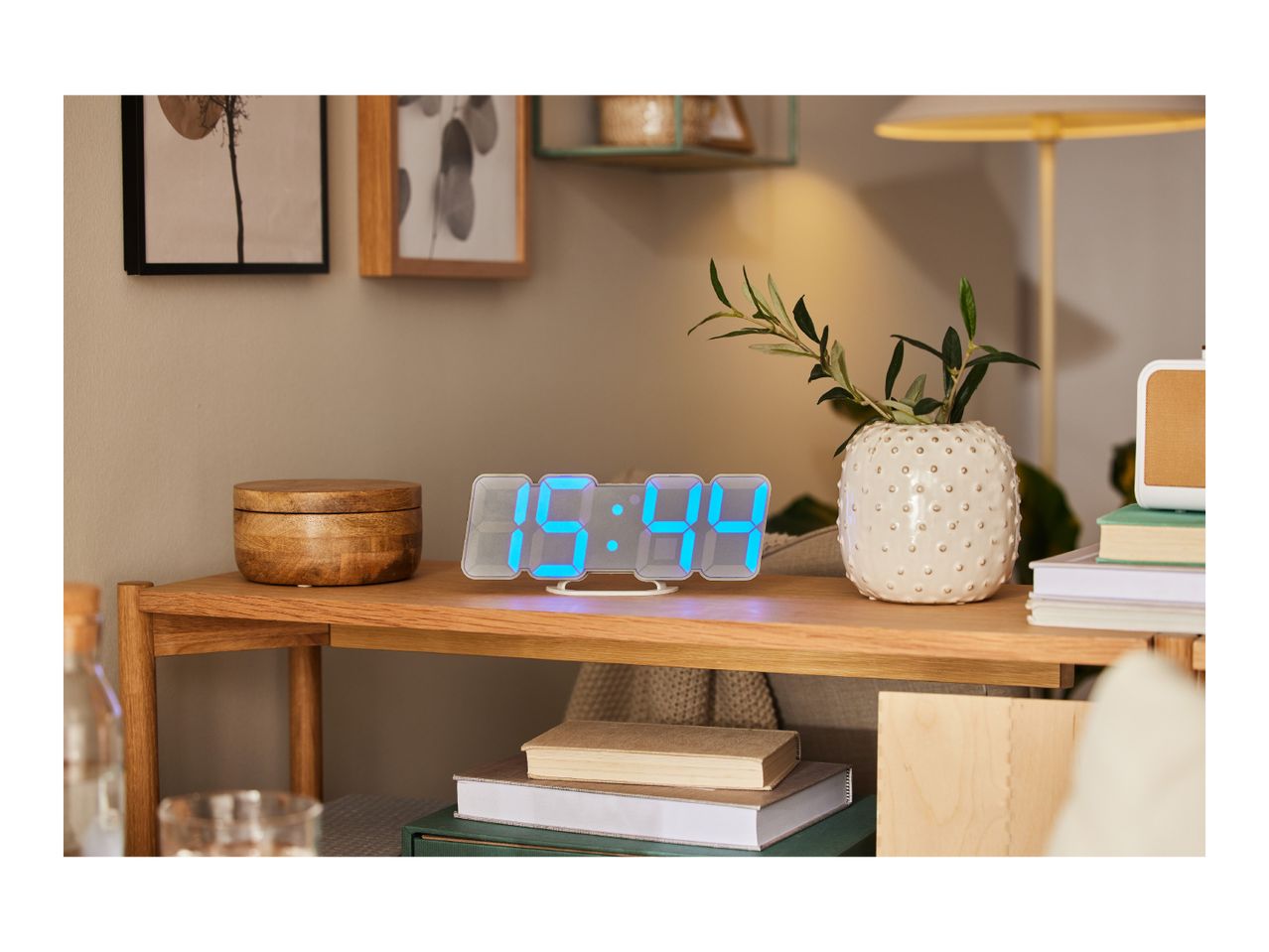 A modern LED digital clock sits on a wooden side table amongst home decor items.