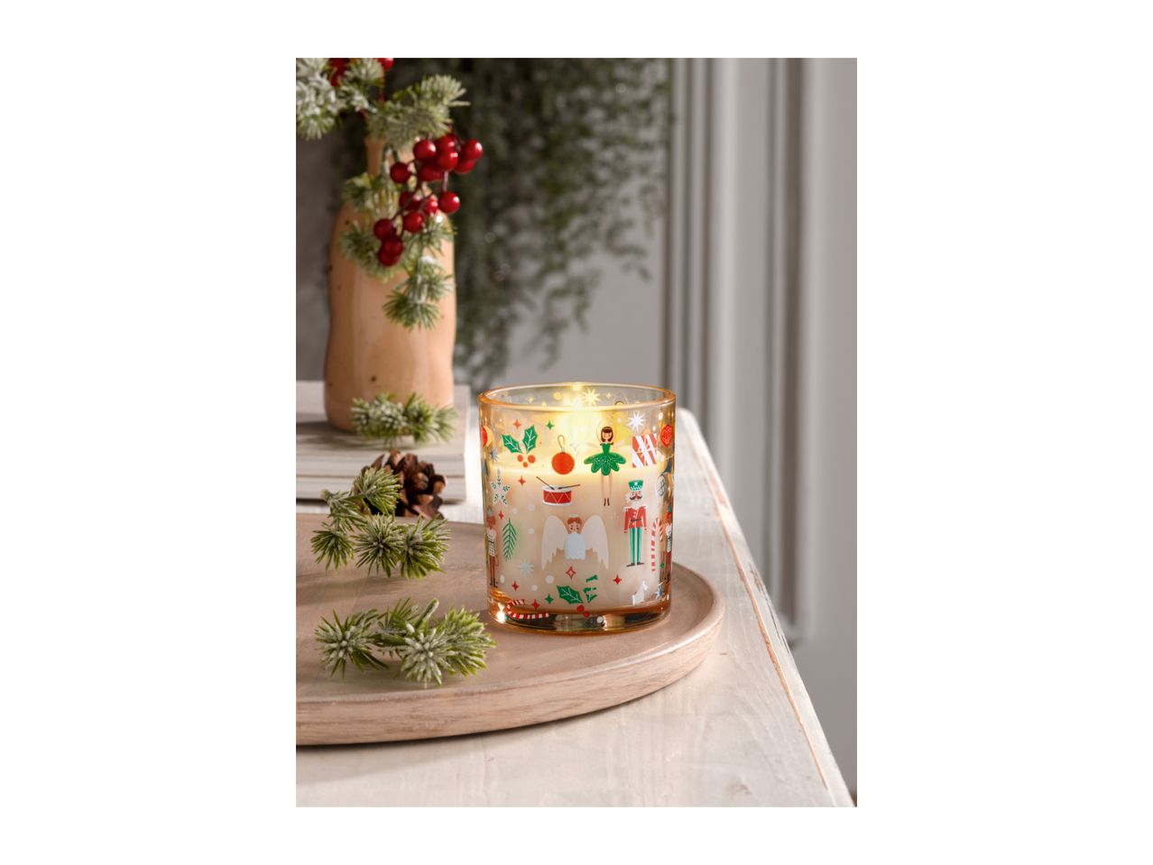 Christmas candle with festive designs, pine branches, and red berries on a wooden tray.