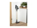 Cordless stick vacuum cleaner mounted on a white wall next to a wooden cabinet with a plant.