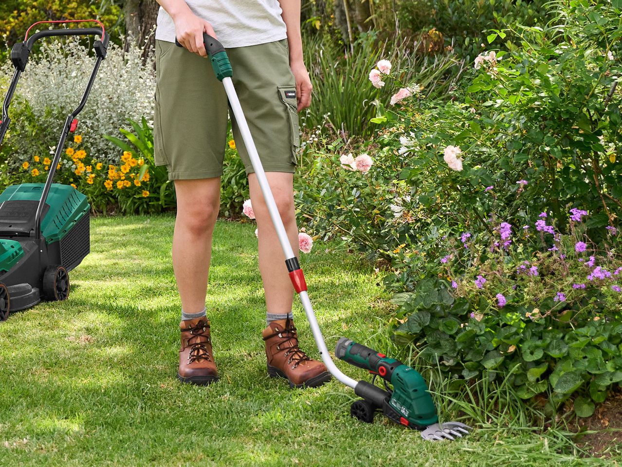 Person using a grass trimmer with wheels to edge a lawn next to a flower bed, with a lawnmower nearby.