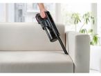 A hand holds a handheld vacuum cleaner cleaning a light beige couch.