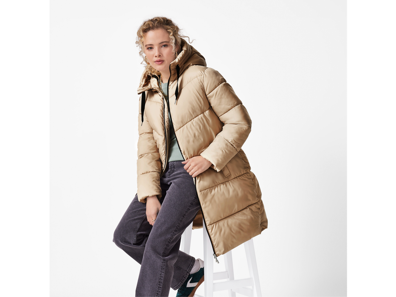 Femeie în geacă bej, blugi gri și adidași verzi, așezată pe un scaun alb.