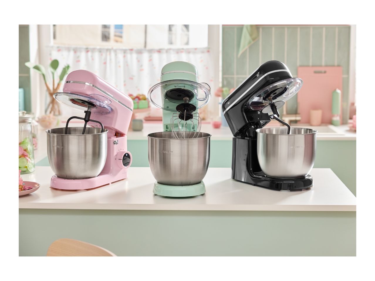 Three stand mixers in pink, mint green, and black on a kitchen counter.