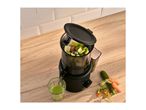 Black juicer with chopped vegetables, a glass of green juice, and whole vegetables on a wooden counter.