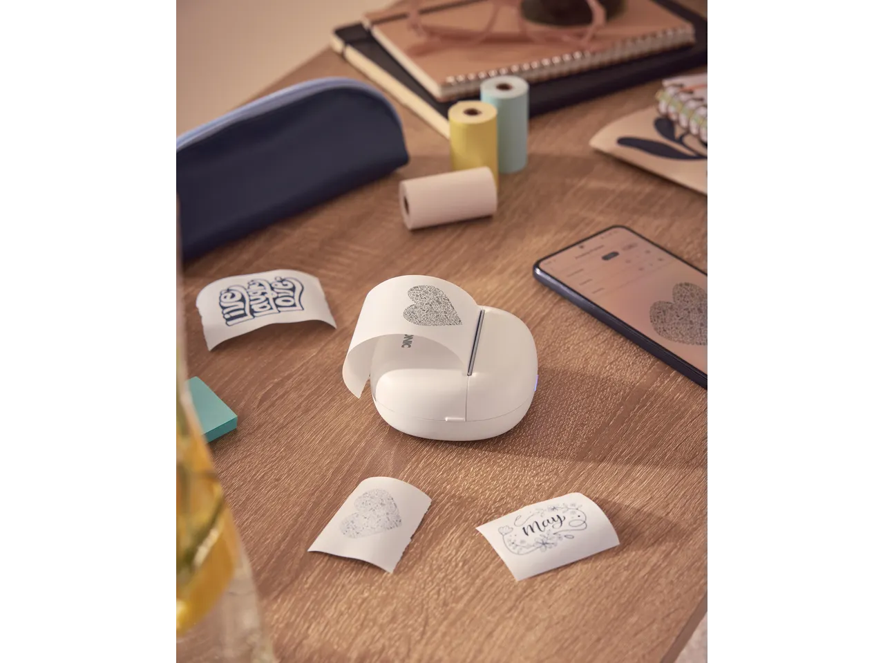 Mini impressora TRONIC imprime um coração e 'May' numa mesa de madeira com outros materiais.
