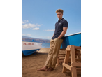 Man in a navy polo shirt and beige trousers leaning on a boat on a sandy beach.