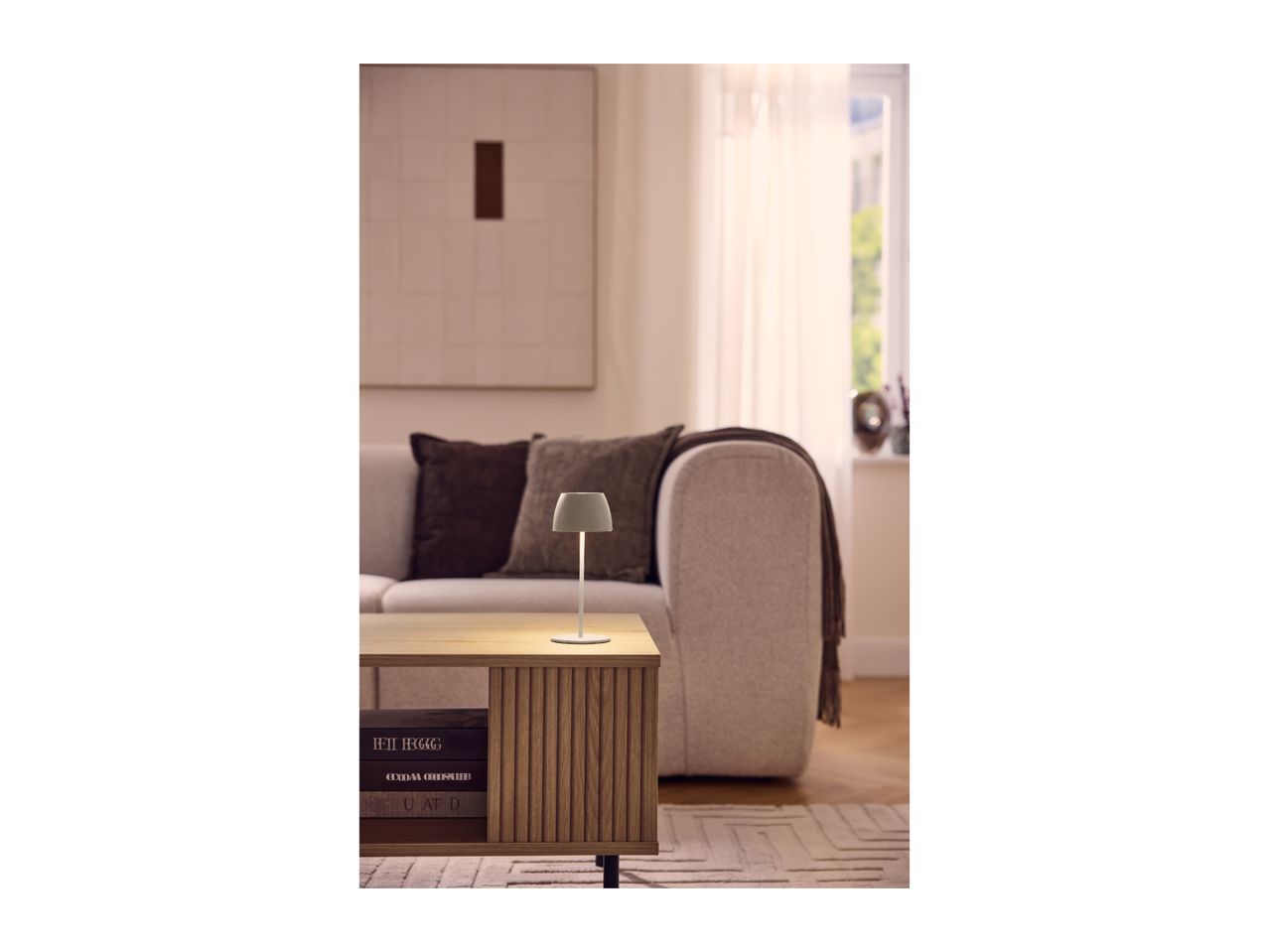 A modern, minimalist table lamp glowing on a wooden coffee table in a cozy living room.