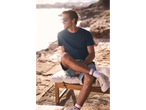 Bărbat relaxat pe o șezlong lângă mare, îmbrăcat cu un tricou bleumarin și pantaloni scurți.