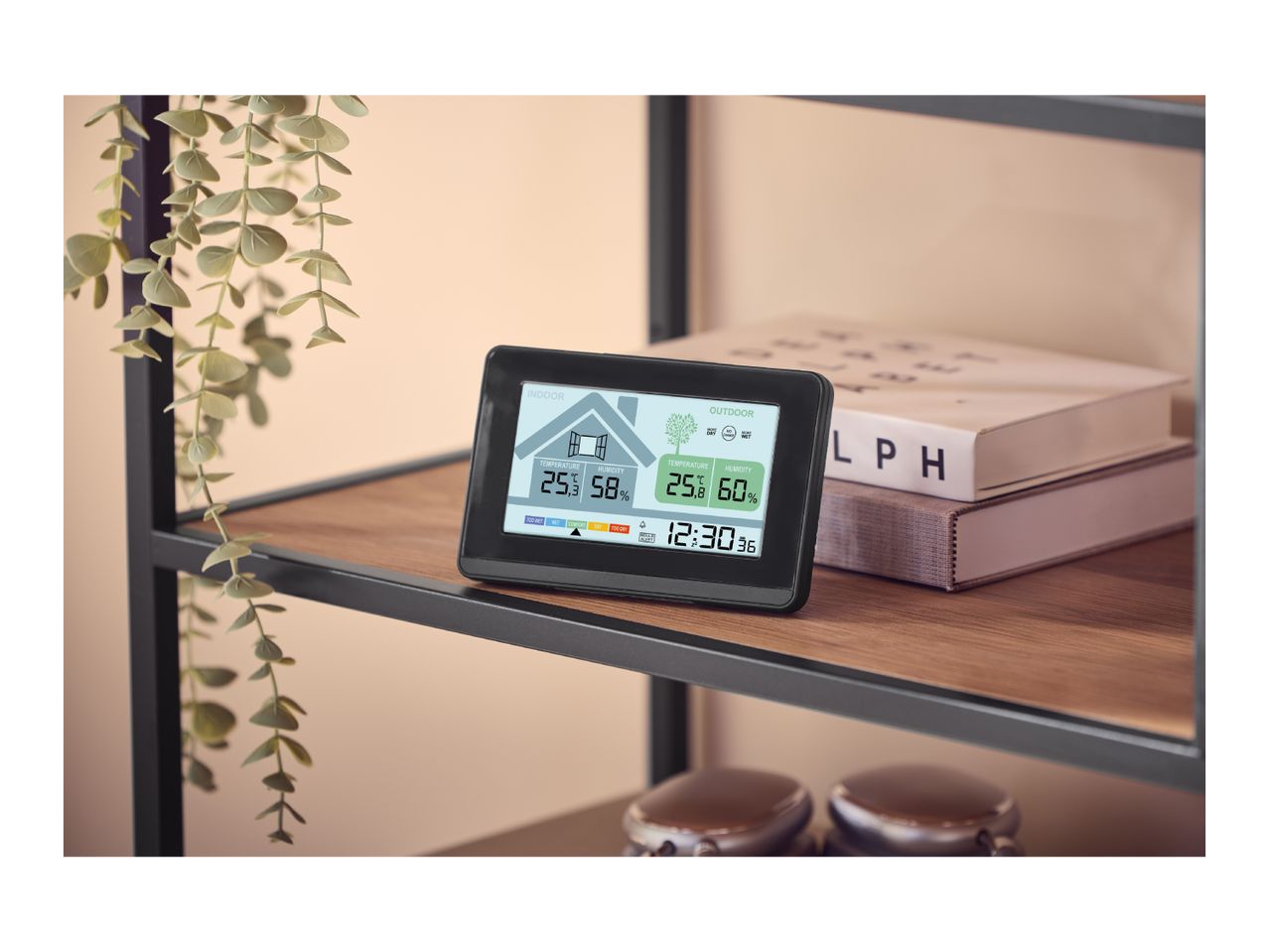 Digital weather station displaying indoor and outdoor temperature and humidity on a shelf with books and a plant.