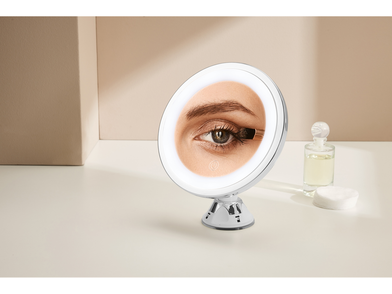 A lighted vanity mirror with a suction cup base sits on a white surface next to cotton rounds and perfume.