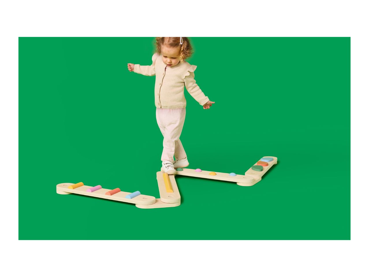 Toddler balancing on a wooden balance beam with colorful steps on a green background.