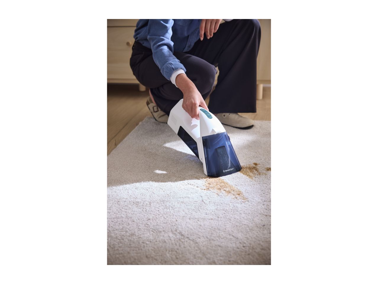 A person cleaning a stain on a light-colored carpet with a handheld carpet cleaner.