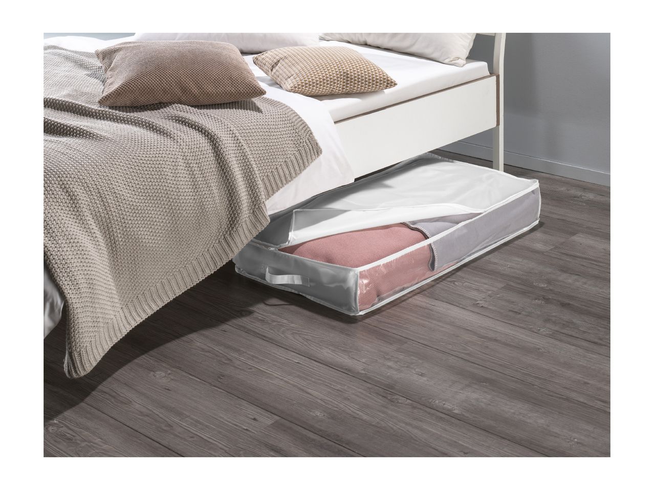 An under-bed storage bag with a clear window, filled with textiles, sits on a wooden floor next to a bed.