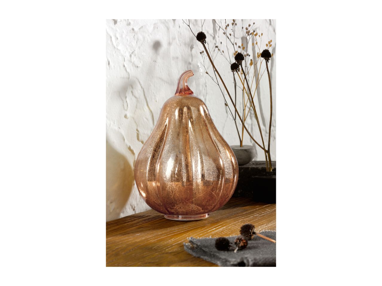A rose gold glass pumpkin decoration on a wooden surface with dried flowers.