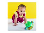 Happy baby in magenta overalls crawling towards a green frog toy.