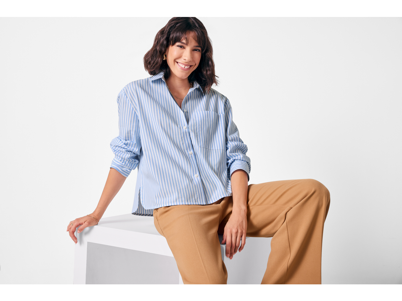 Mulher com camisa às riscas azuis e brancas e calças castanhas, sentada e a sorrir.