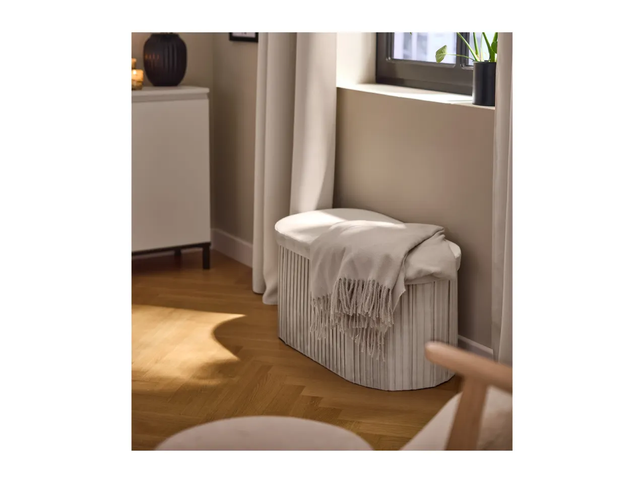 A light gray storage ottoman with a blanket on top, next to a window and a white cabinet.