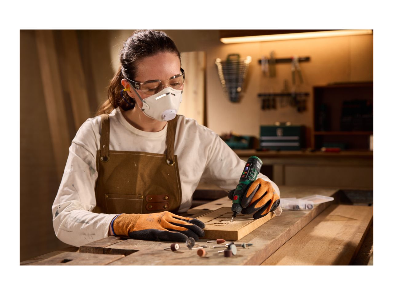 Woman in safety gear using a rotary tool to engrave wood, with various bits on the workbench.