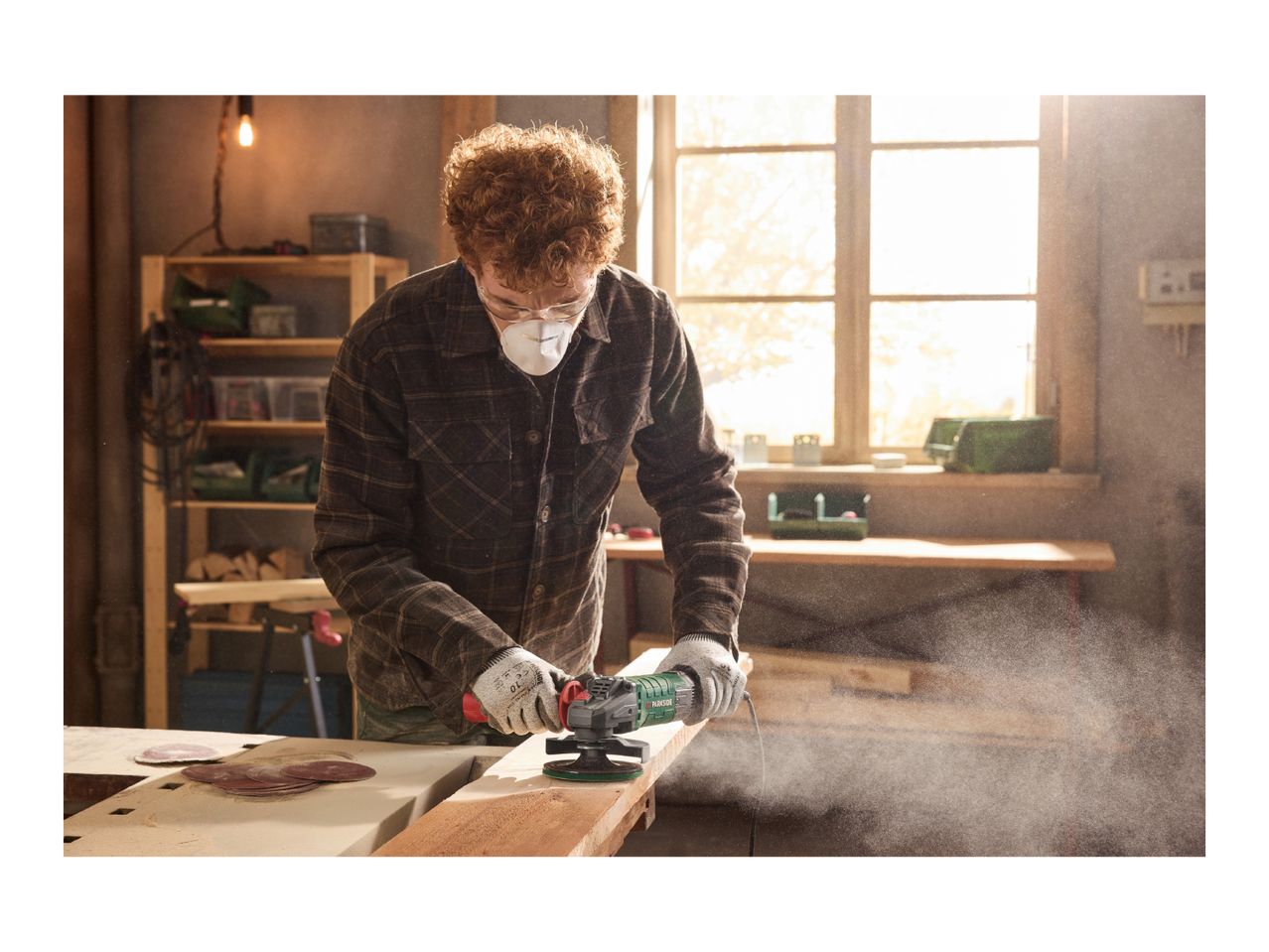 Man in workshop sanding wood with an angle grinder, wearing a mask and safety glasses.