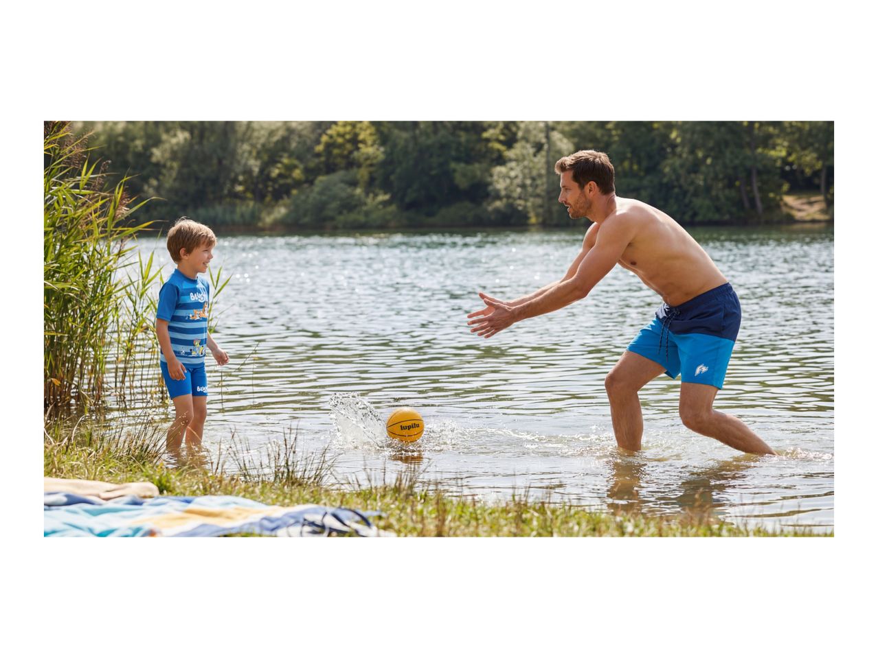 Father and son playing with a ball in the lake, wearing swim shorts and a rash guard.