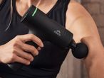 A person in a black sports top uses a black massage gun on their arm.