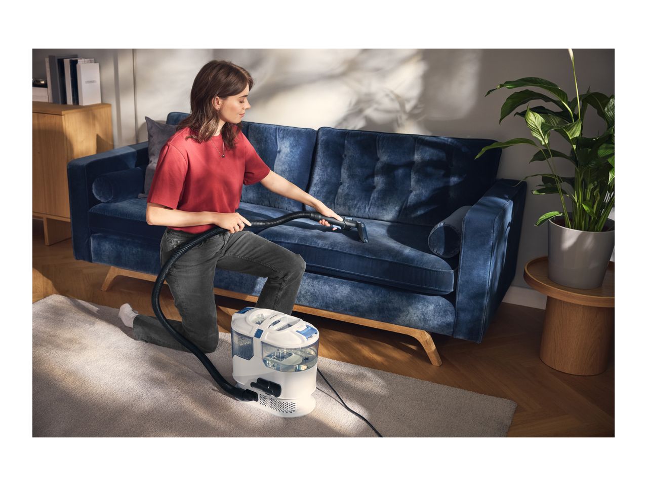 A woman cleans a blue velvet sofa with a portable spot carpet cleaner.