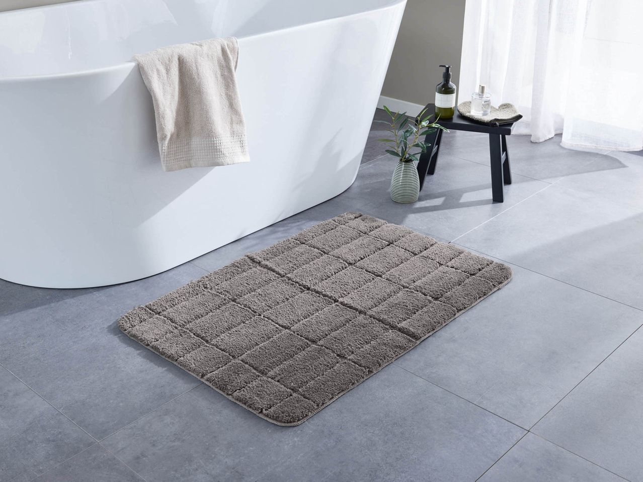 Un tappetino da bagno grigio con motivo a blocchi su un pavimento piastrellato, accanto a una vasca da bagno bianca.