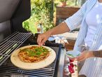 Nő pizzát tesz egy grillen lévő pizzakőre, friss rukkolával díszítve.