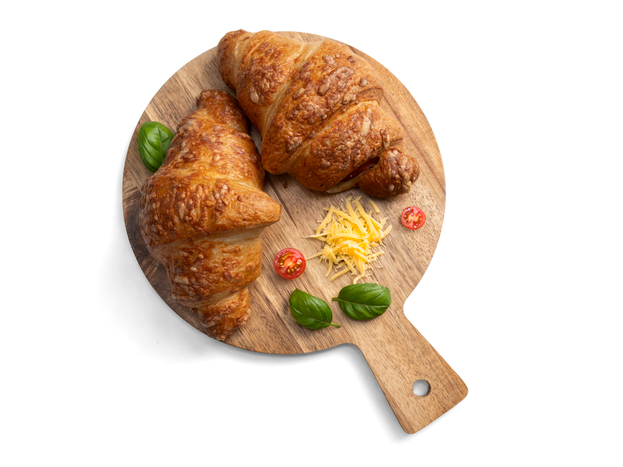 Két sajtos-sonkás croissant egy fa vágódeszkán bazsalikommal, koktélparadicsommal és reszelt sajttal.
