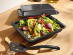 Roasted vegetables in a black baking dish with a matching lid and spatula on a wooden board.