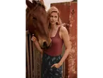 Femeie cu un top maro și pantaloni cu model lângă un cal brun.