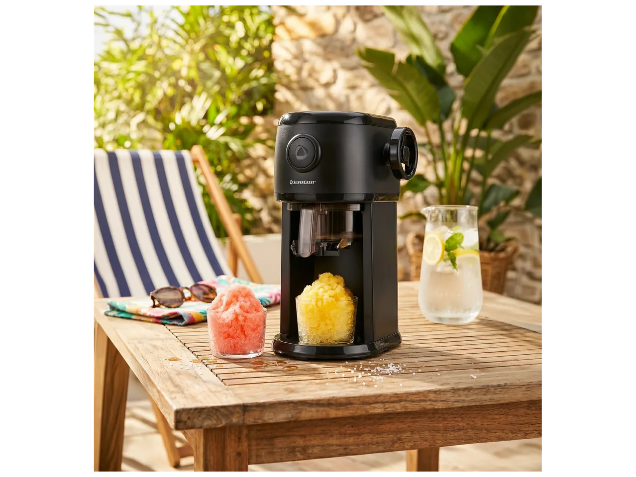 Machine à glace pilée noire avec deux verres de granité coloré sur une table en bois en extérieur.