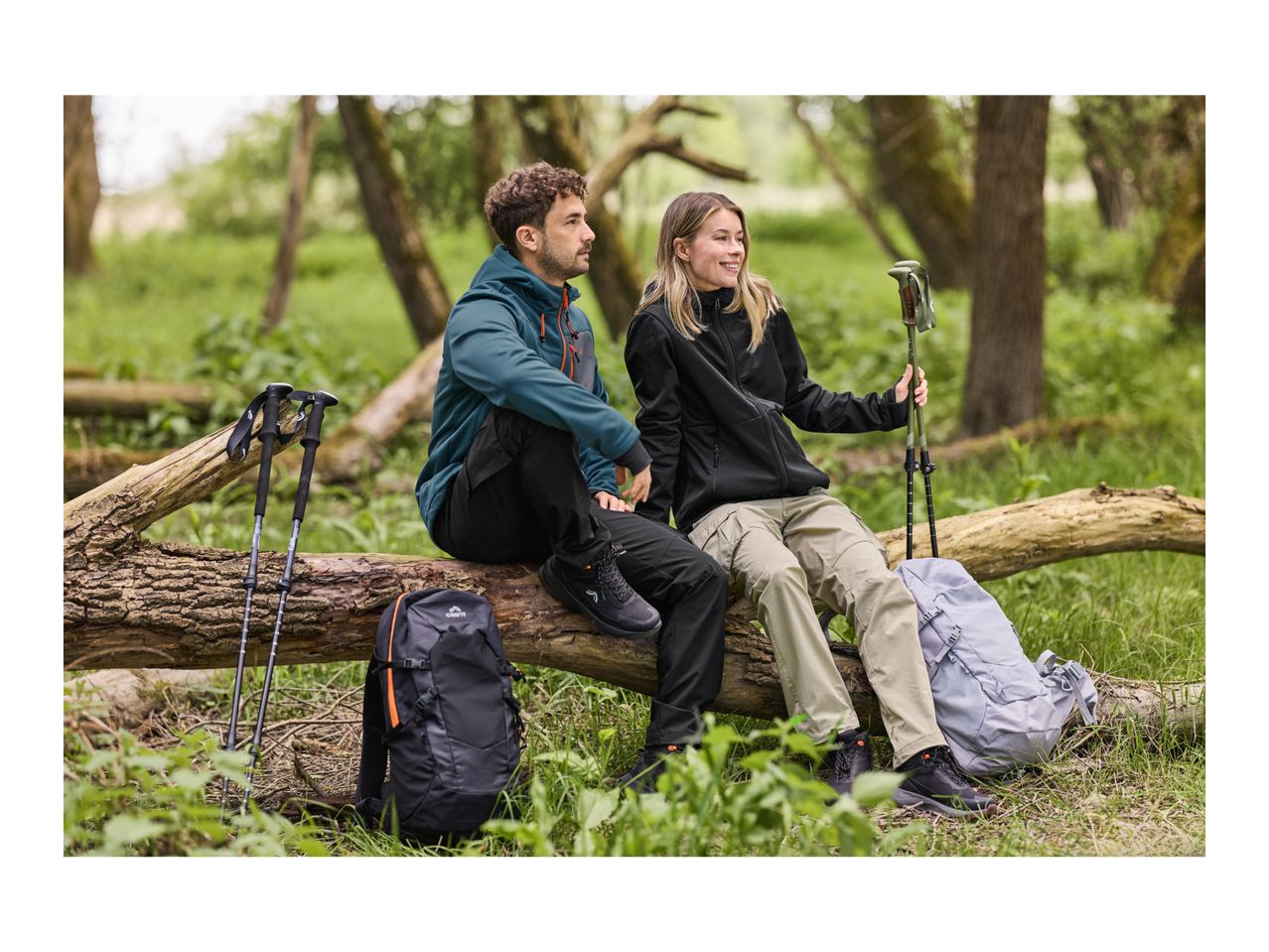 Two hikers in outdoor clothing and shoes, with backpacks and walking poles, sitting on a log.