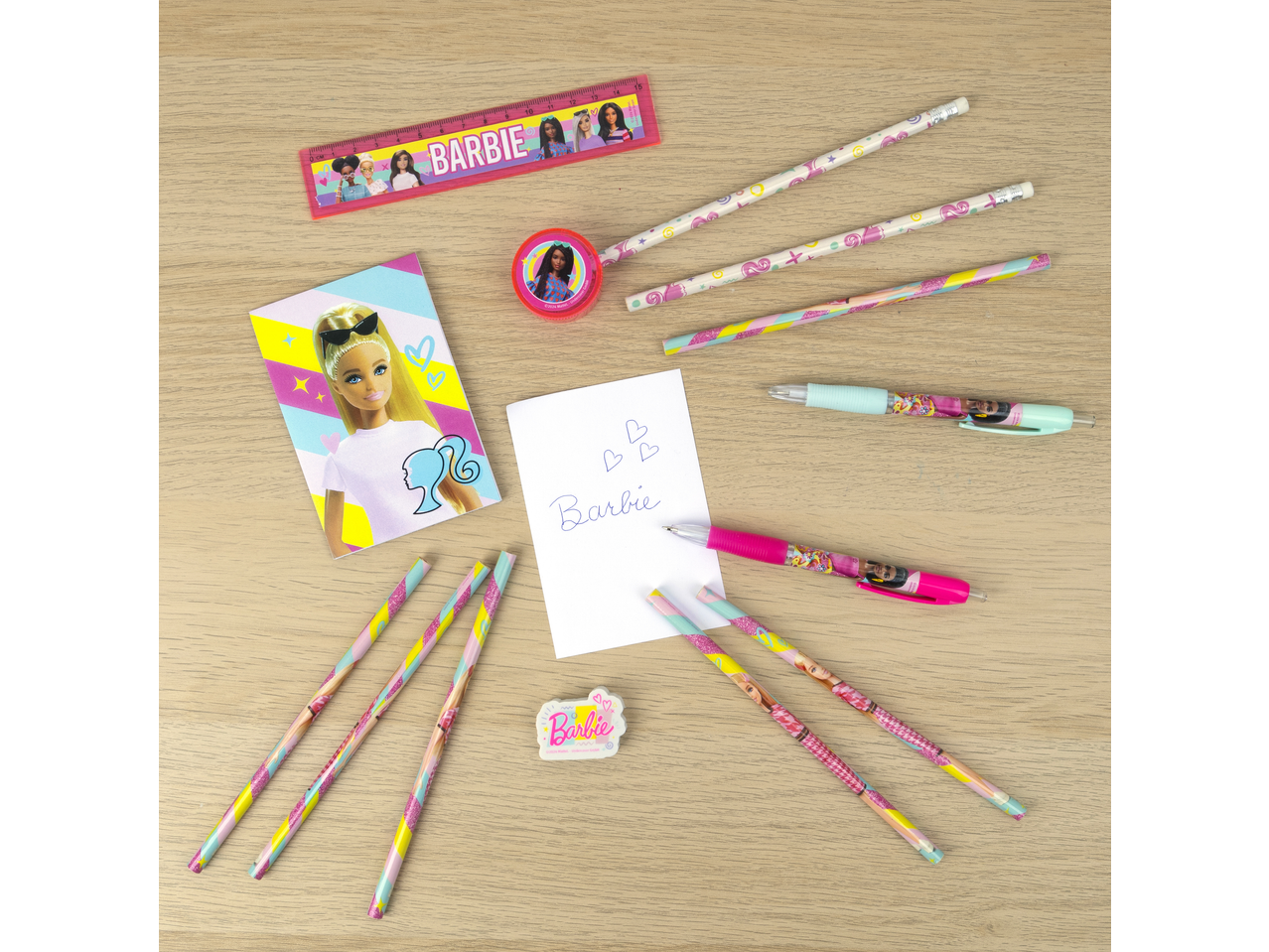 Conjunto escolar Barbie com régua, lápis, afia, canetas e borracha numa mesa de madeira.