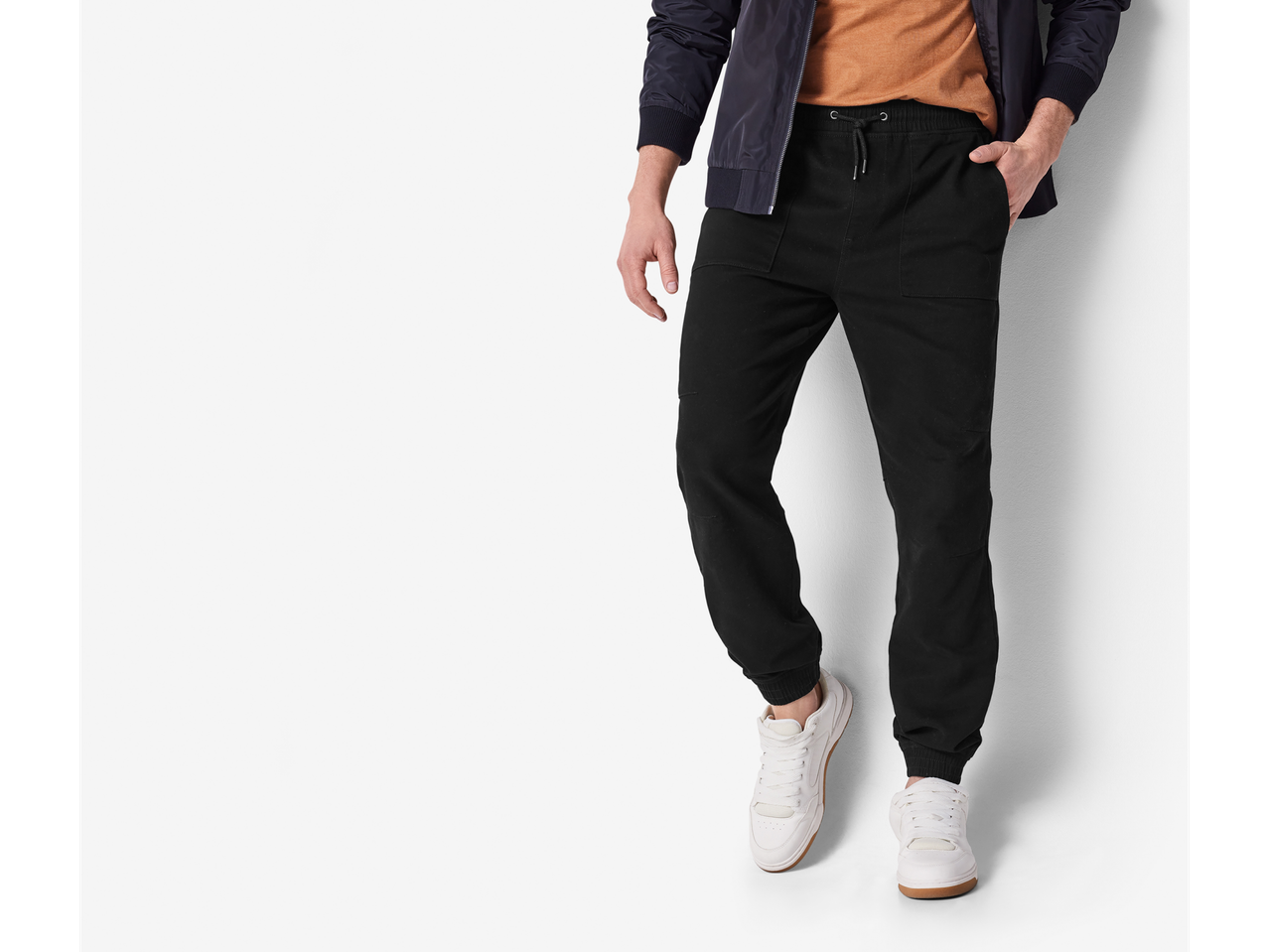 A man wearing a navy bomber jacket, an orange t-shirt, black joggers, and white sneakers.