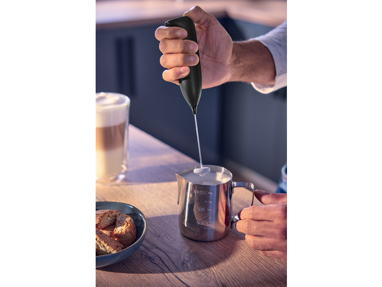 Hand frothing milk in a stainless steel jug with a handheld frother, next to a latte and biscotti.