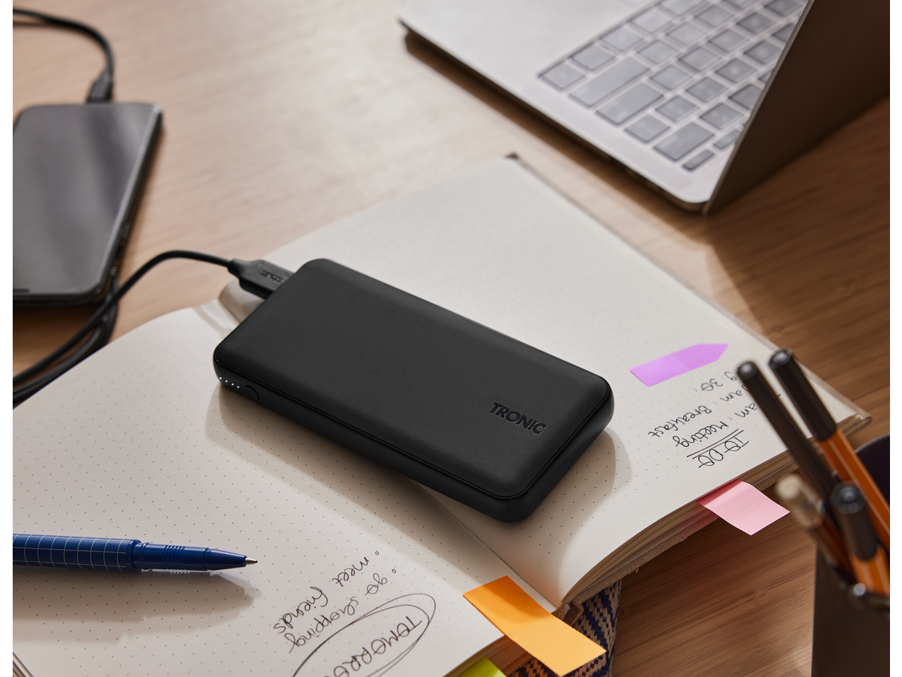 TRONIC 10000 mAh Power Bank charging a phone on a desk with a notebook and laptop.