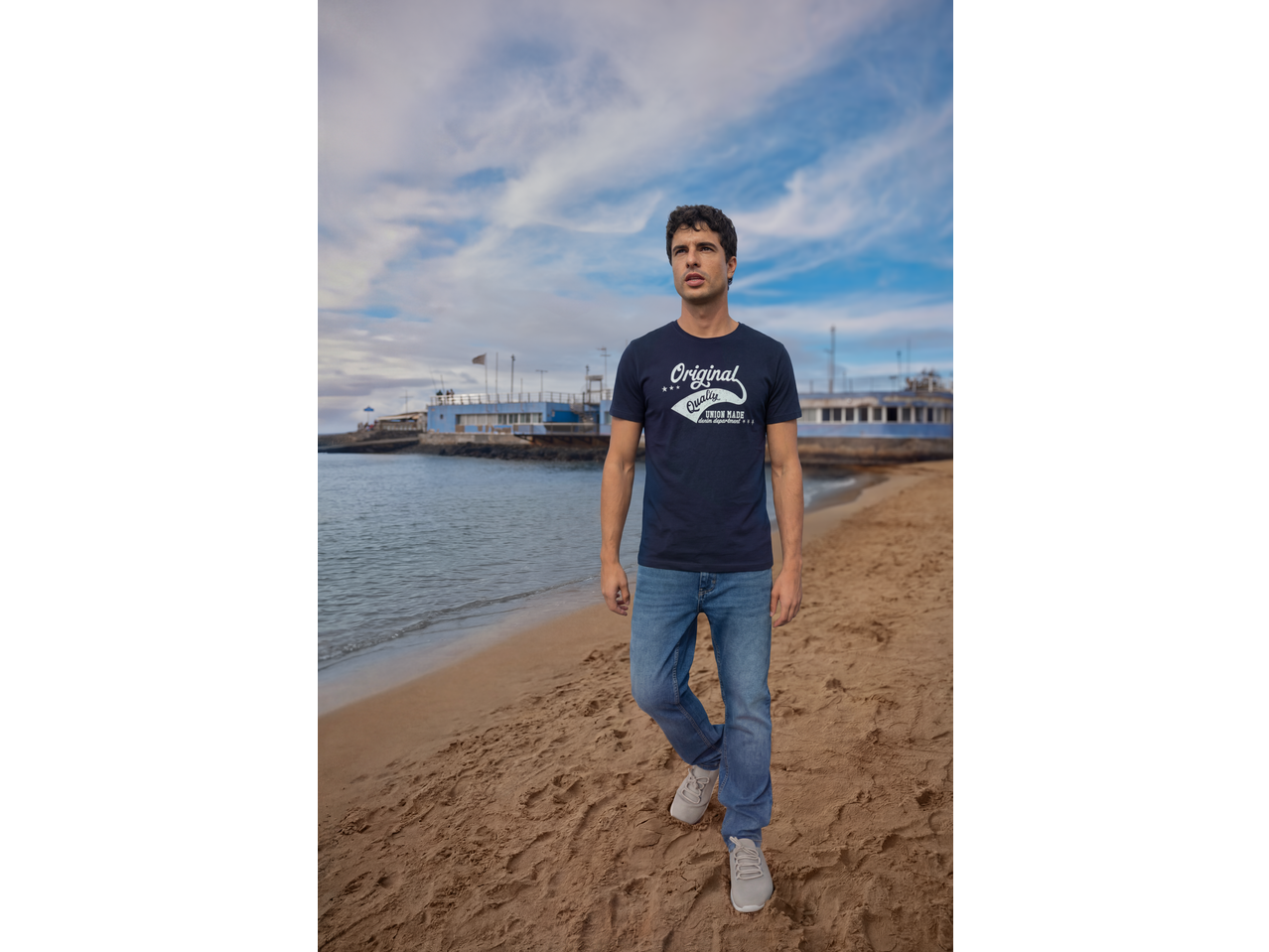 Άνδρας με σκούρο μπλε μπλουζάκι 'Original Quality' και τζιν περπατά στην παραλία.