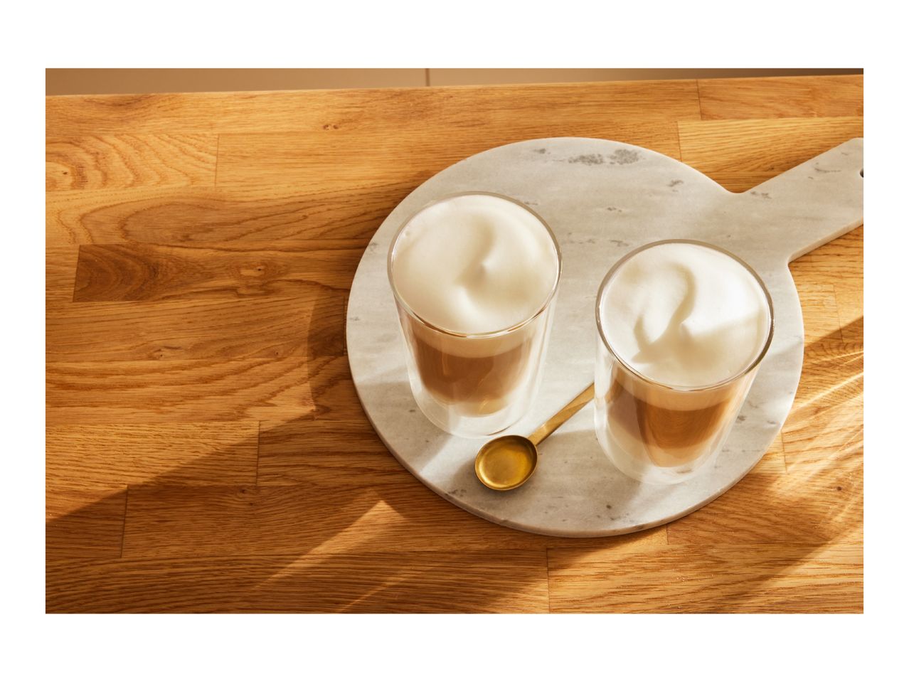 Two glasses of latte macchiato with foamed milk on a marble board and wooden table.