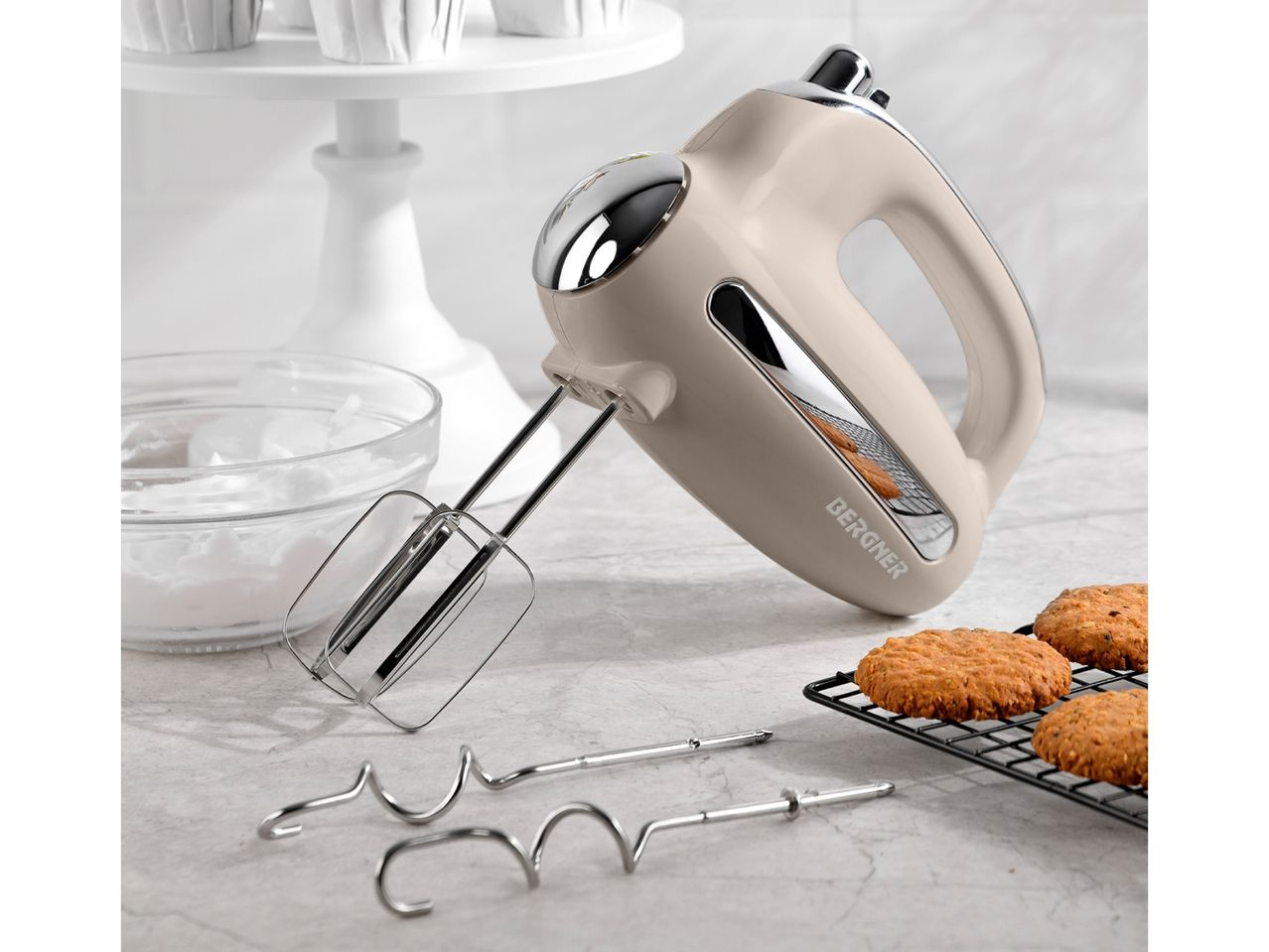 A beige retro-style hand mixer with beaters and dough hooks sits on a countertop next to freshly baked cookies.