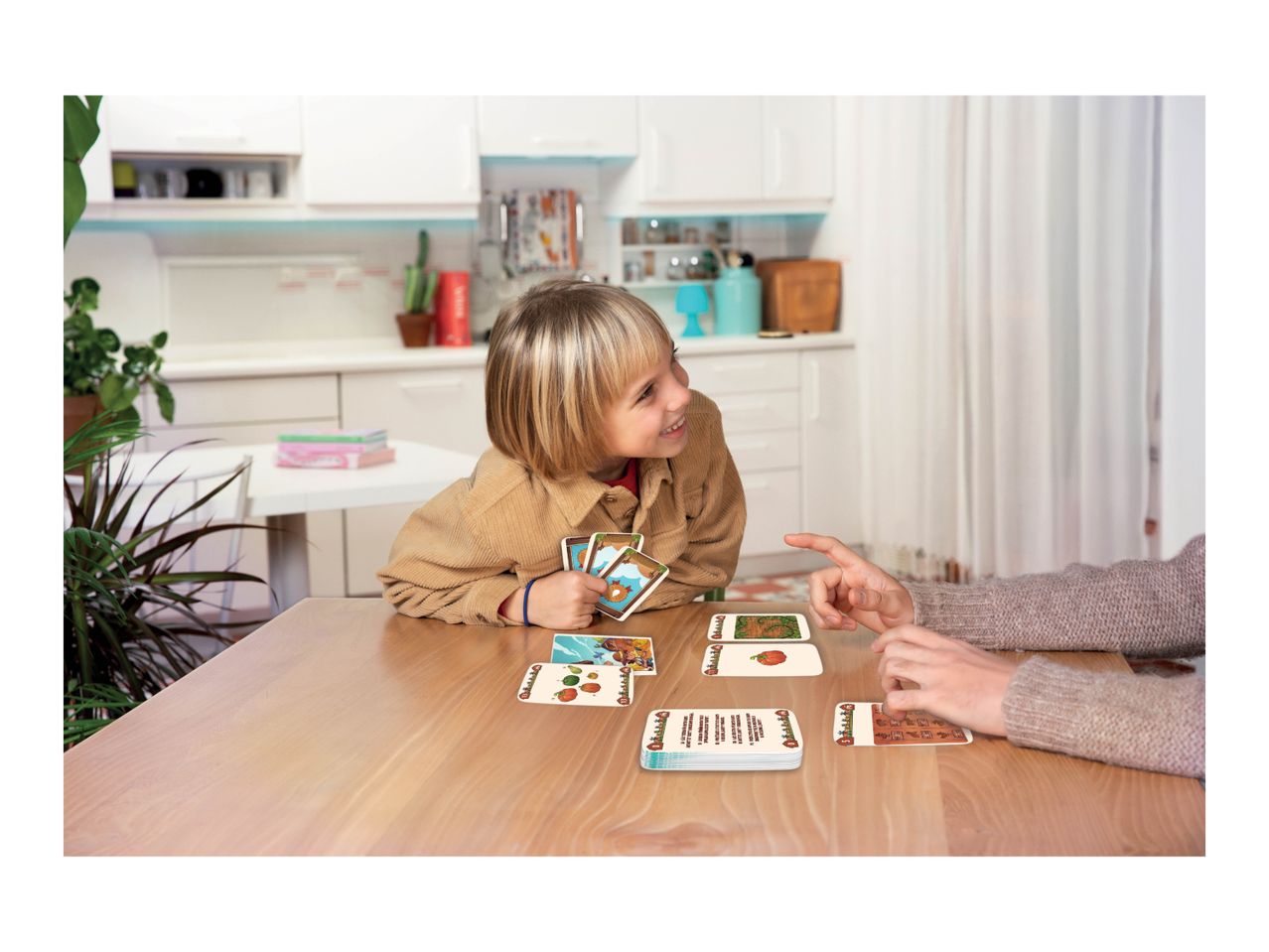 Child and adult playing a card game at a wooden table in a kitchen.