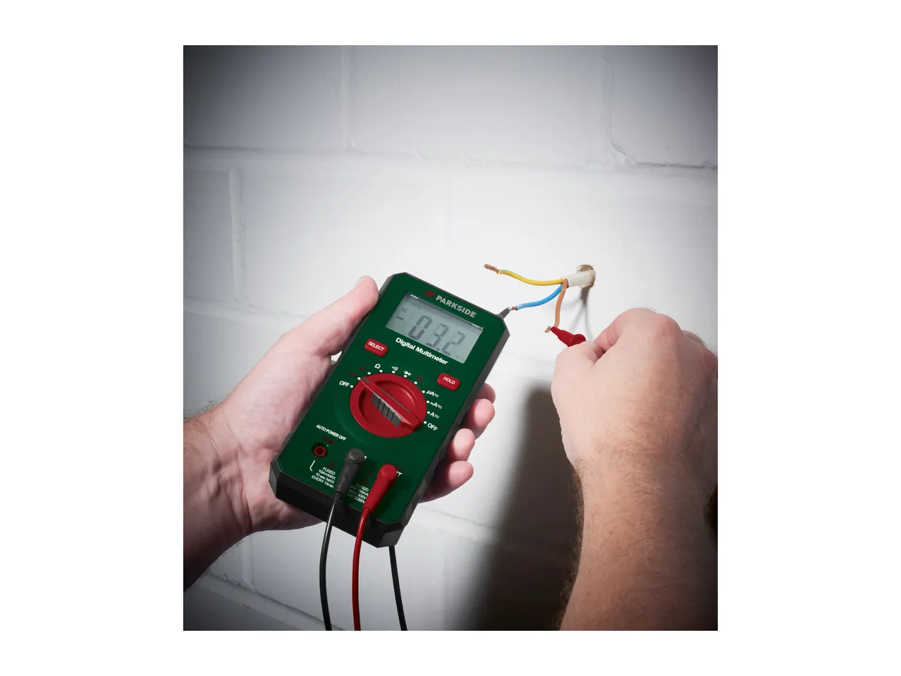 A person's hands using a digital multimeter to test electrical wires coming out of a wall.