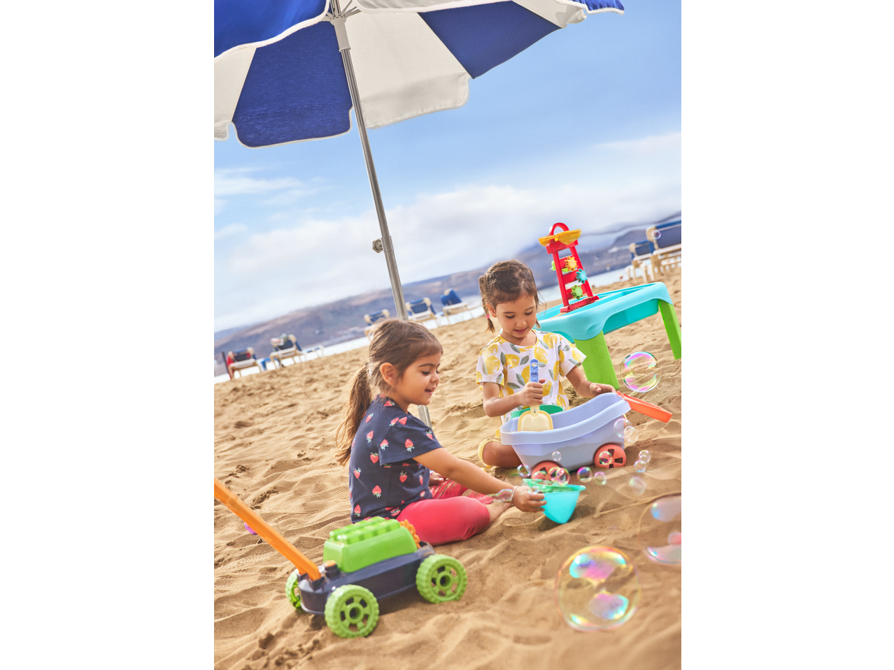 Two children playing with a Lupilu Sand and Water Activity Table and other toys on a sandy beach.