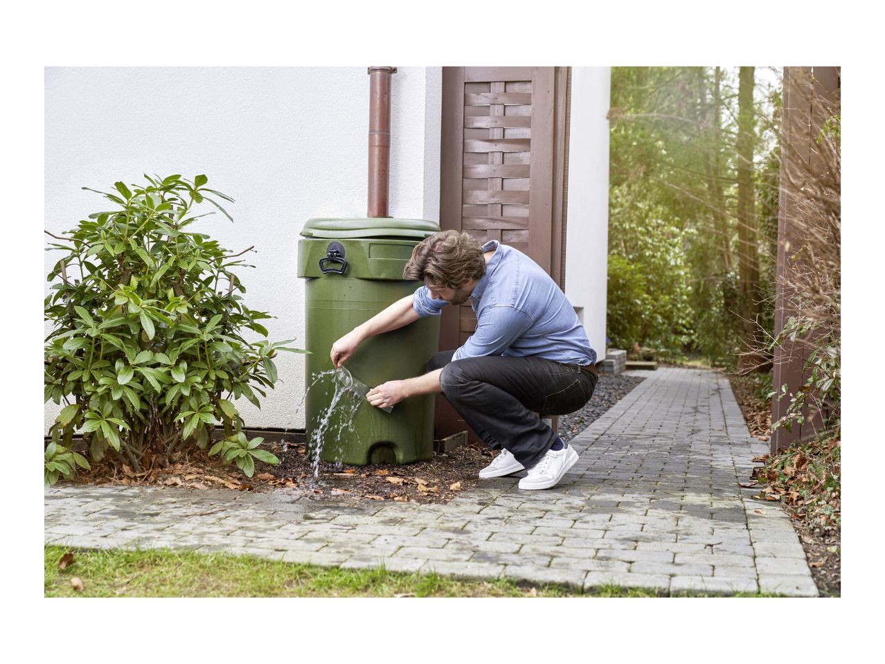 Man collecting rainwater from a green rain barrel with a downspout connection.