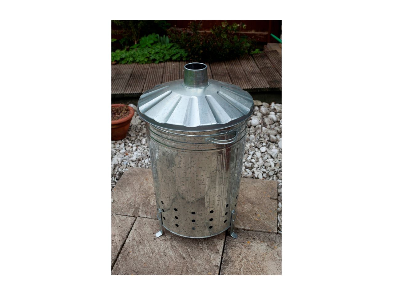 Galvanized steel garden incinerator bin with lid, on a patio with gravel and plants.